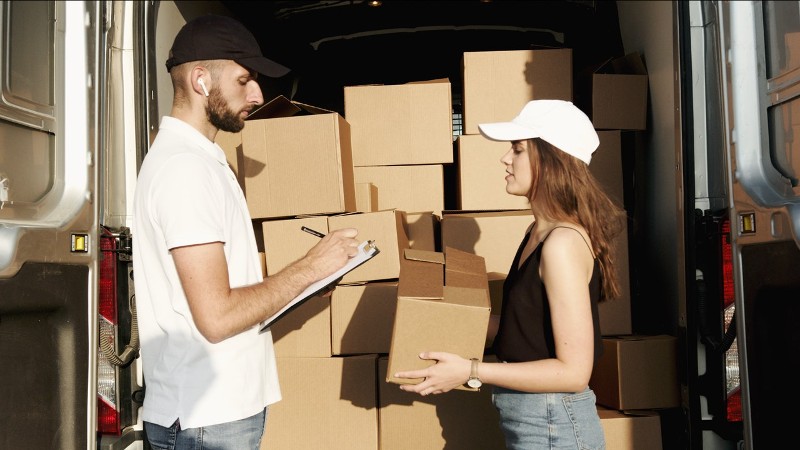 Empleado verificando el seguro de mercancías a su clienta mientras de fondeo hay varias cajas cafés de mercancía adentro de camioneta